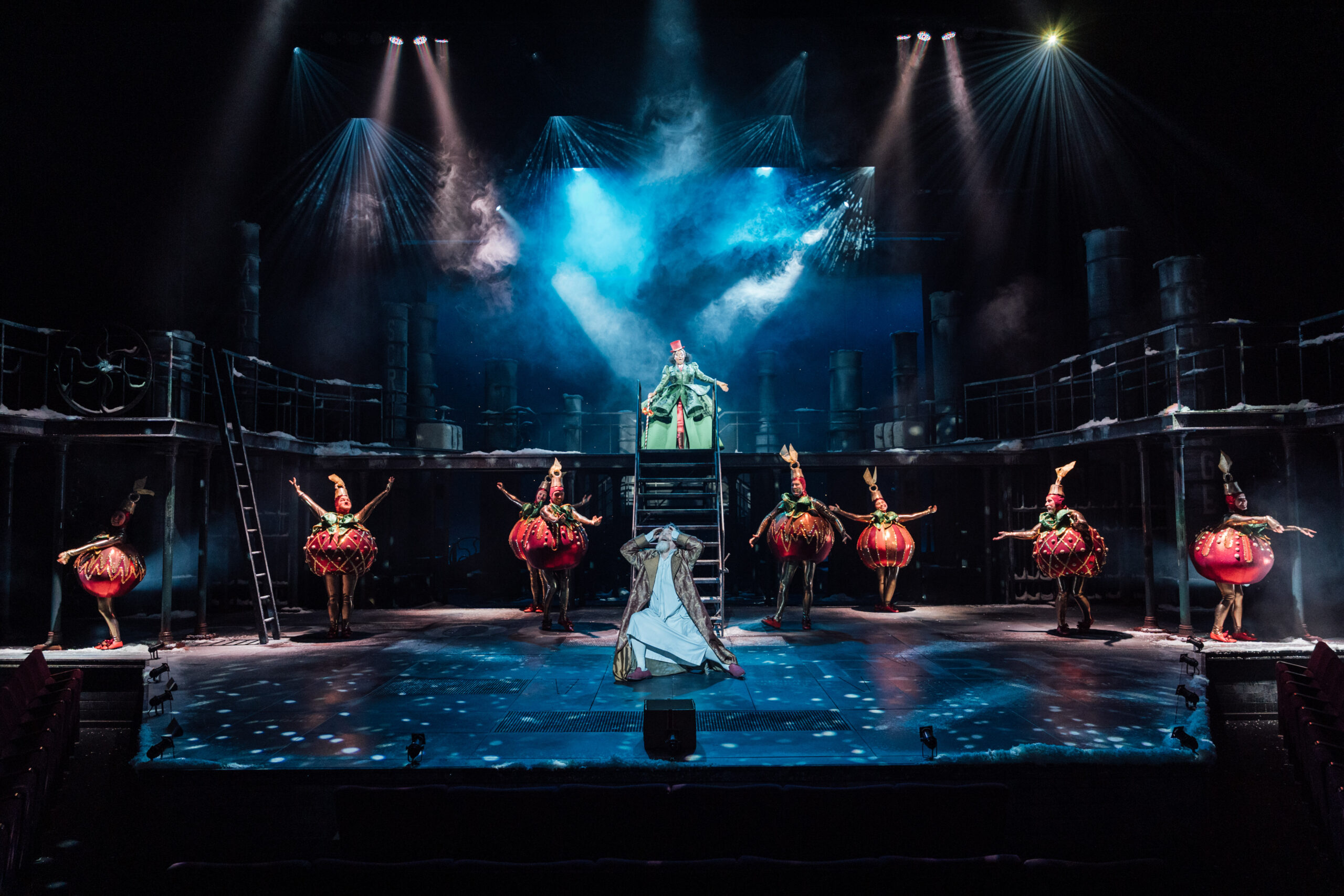 a large stage with several actors dressed as christmas baubles, a man in the middle in a nightgown and a woman at the top of some steps in a dress under a spotlight