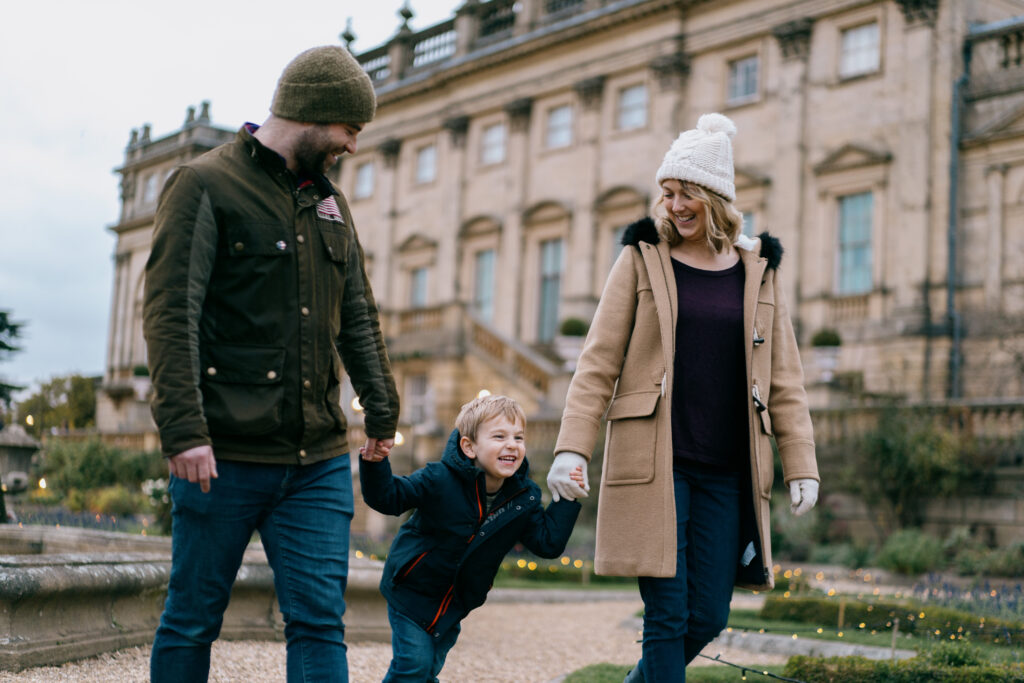 a pair of adults holding hands with a young child in a manicured garden in front of a large stately home