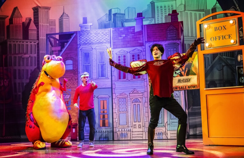 a theatre stage with an actor in front and one behind next to a dinosaur