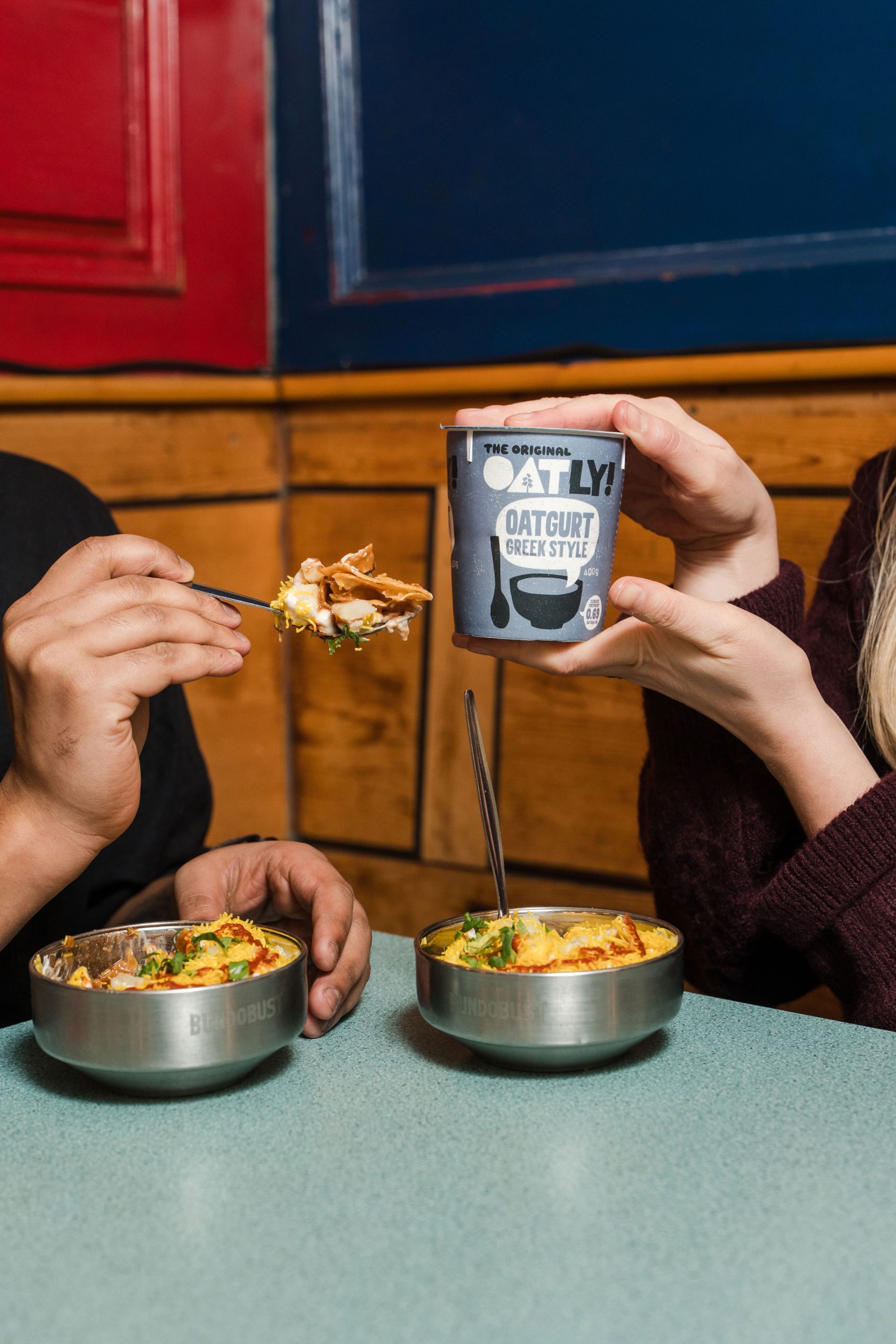 two people sat at a table with bowls of food in front of them, one holding a fork with food on and the other holding up a pot of oatly branded producted