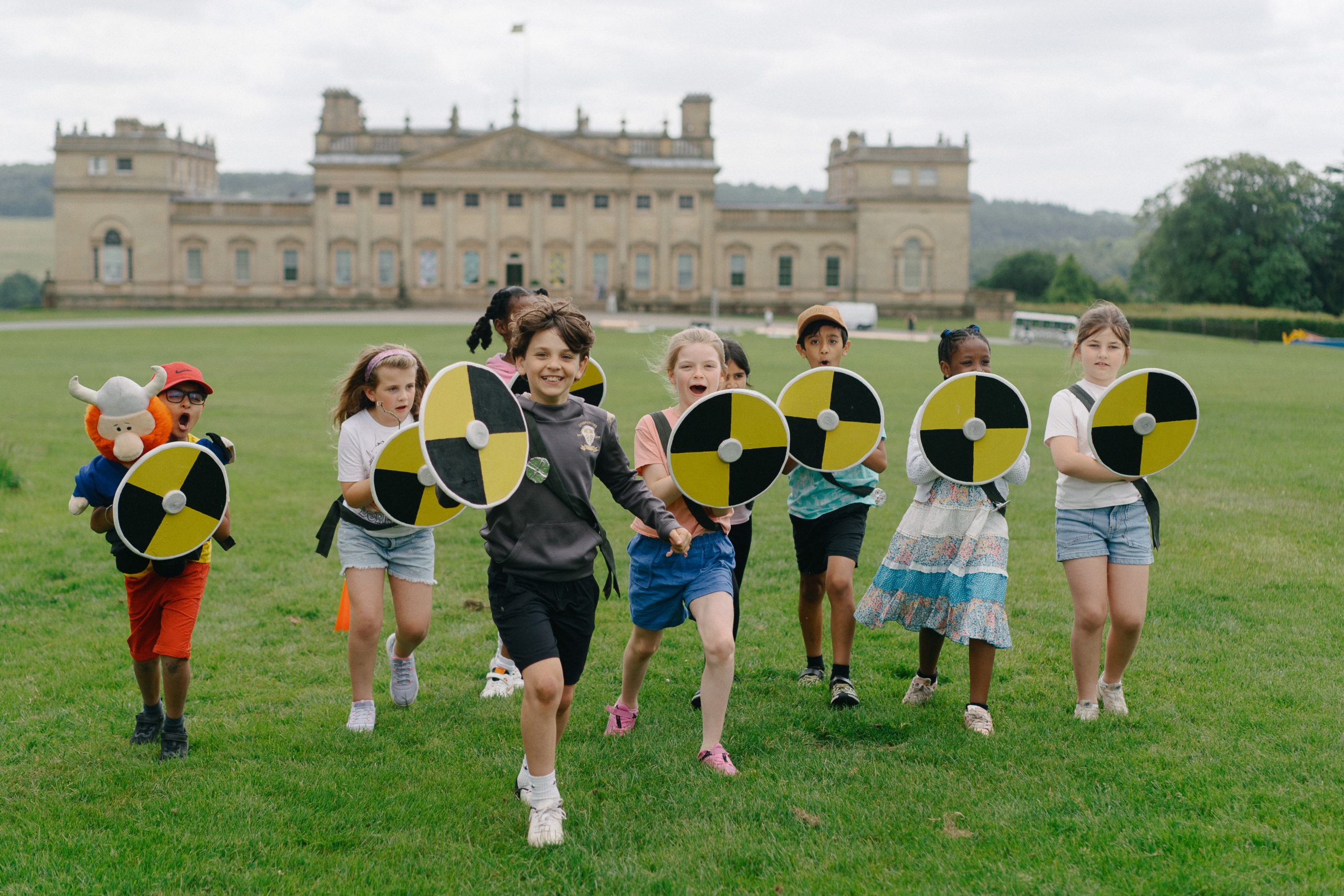 a group of children running towards the camera holding round shields with a large historic stately home in the background
