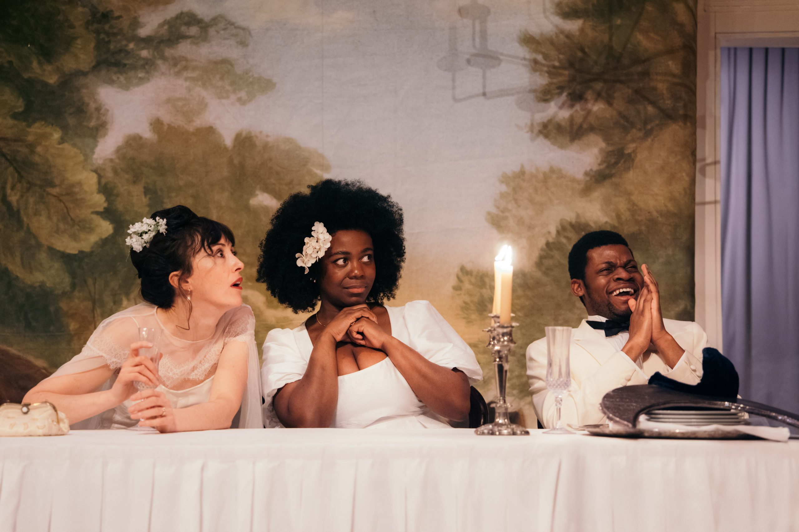 three people on a theatre stage sit at a long table with candles and a white tablecloth on top, and a backdrop resembling a forest