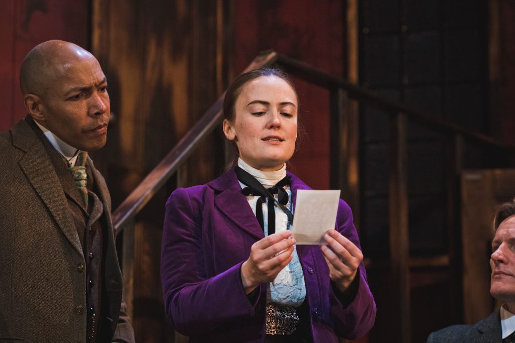 a woman in old victorian dress looking at a piece of paper whilst someone looks over her shoulder