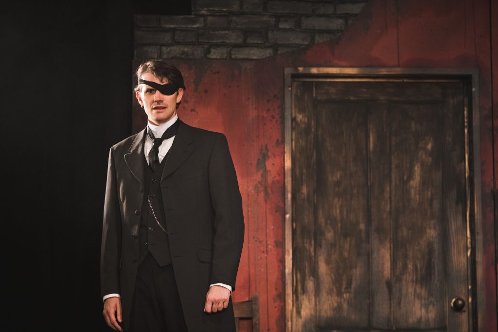 a man with an eye patch wearing an old victorian suit stood alone in a room