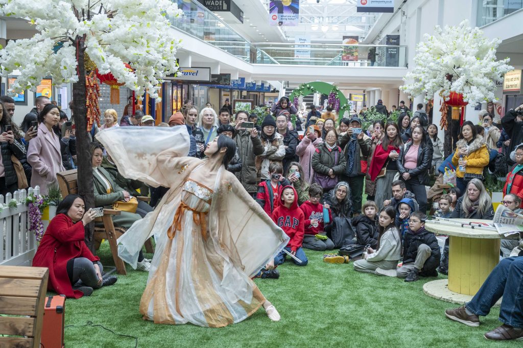 a lady in a traditional chinese dress with large sleeves performing a dance in front a crowd of people in the middle of a shopping centre