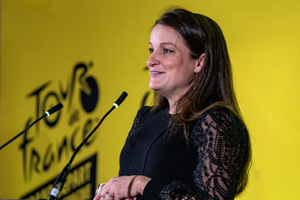 a woman at podium speaking with yellow background and the tour de france logo