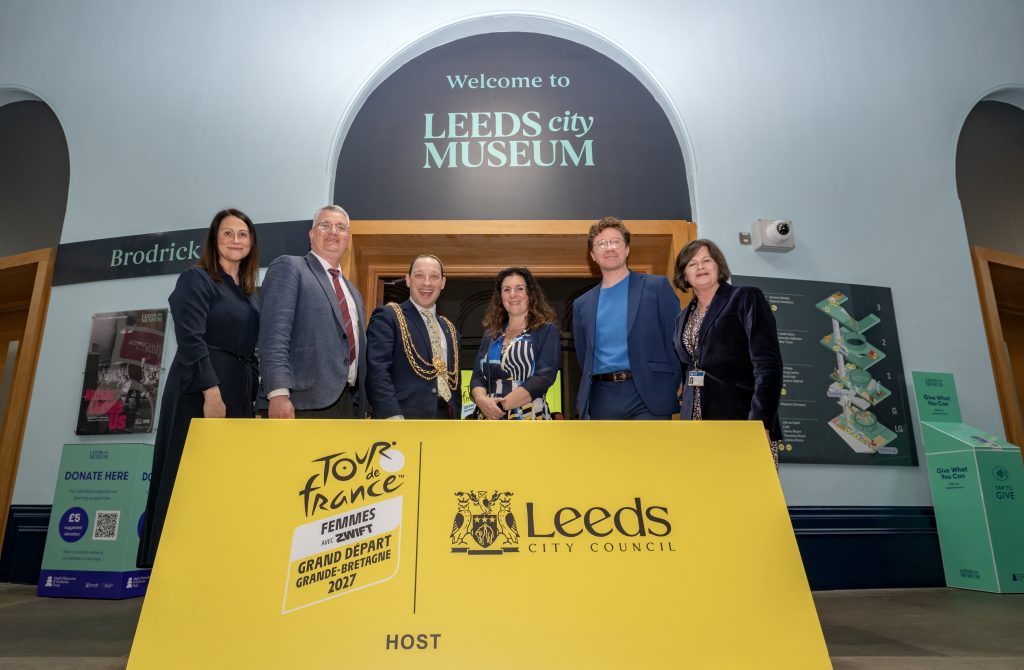 six people, mix of genders standing at a podium with the Leeds City council and Tour de France logos in bright yellow