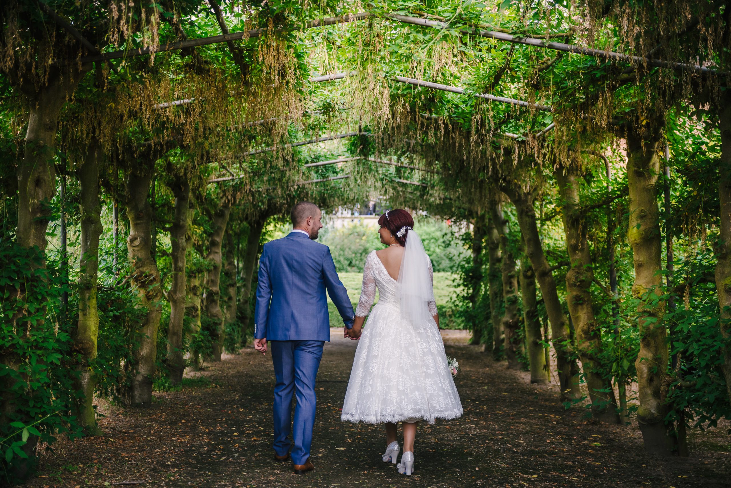 a wedding couple, in a suit and bridal dress, walking amongst a floral archway