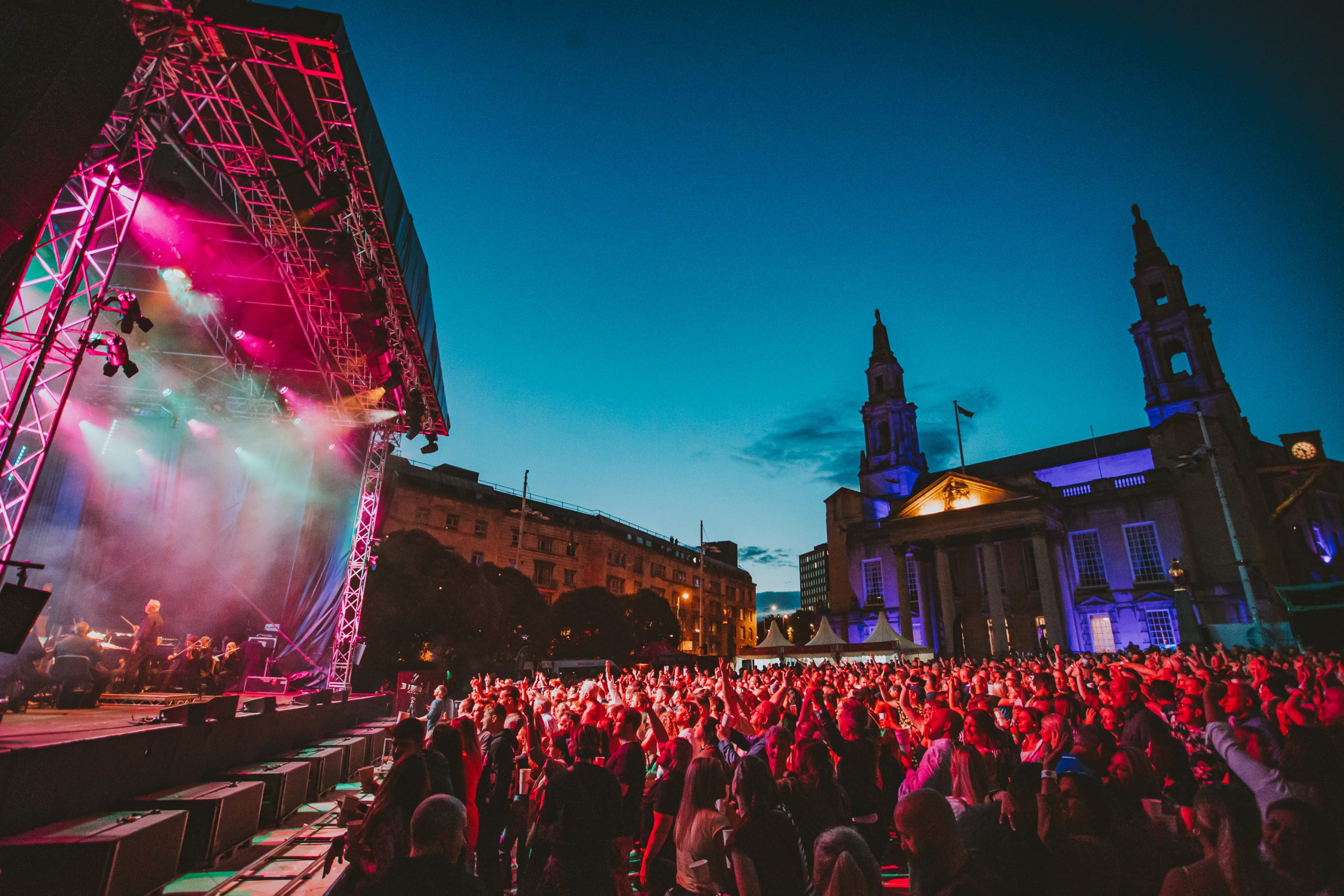 an outdoor stage at nighttime with colourful lights, a large crowd