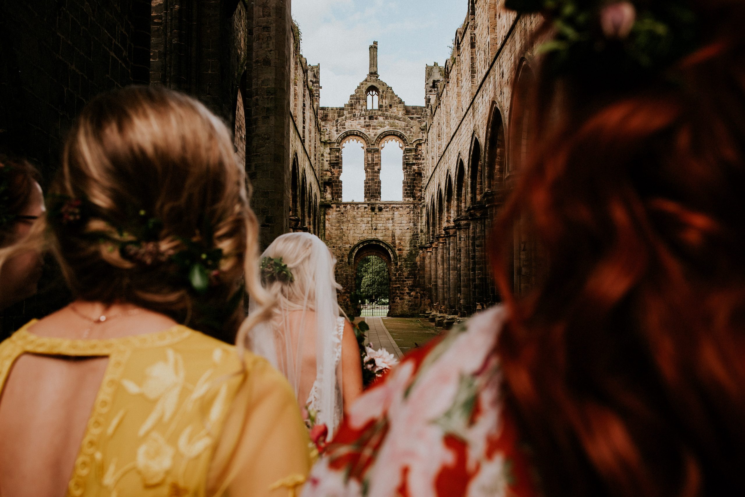 a group of people in nice dresses, with a bride in a veil ahead, in the ruins of an old building