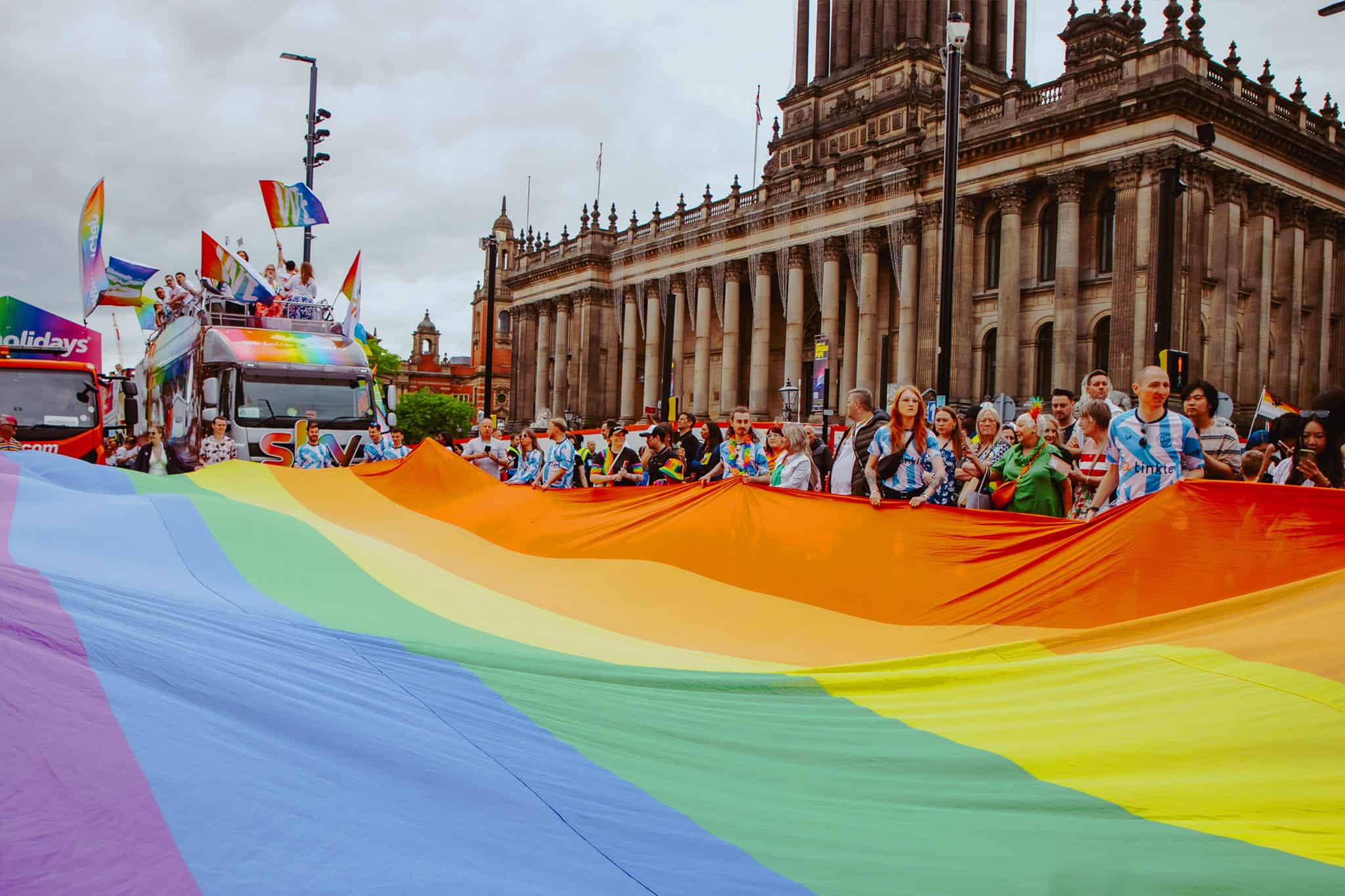 a very large pride flag being held on a street by many people in front of a large historic town hall