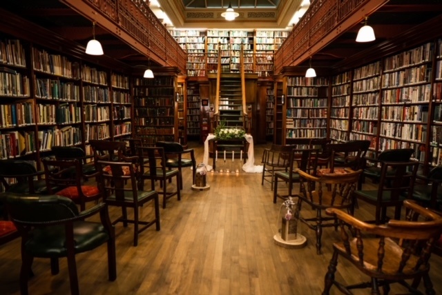 a library with book shelves lining the walls, a second floor, and chairs lined up facing an archway decorated with flowers 