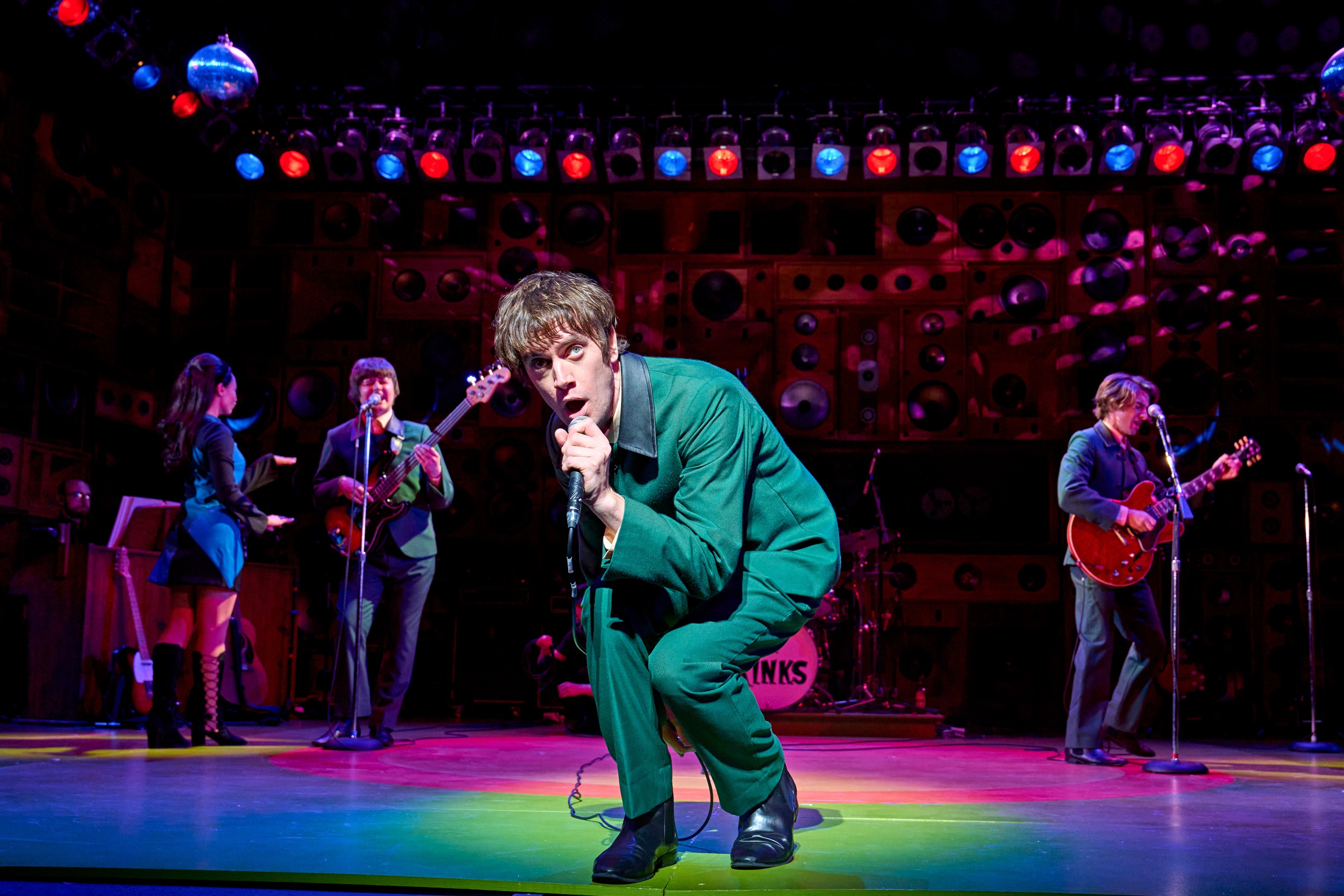 a theatre stage, with someone stood at the front singing into a microphone in a spotlight, and a band playing behind in lower light