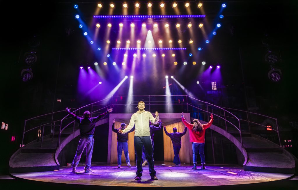 a theatre stage, five actors under some colourful lights with one in a spotlight at the front looking at the audience