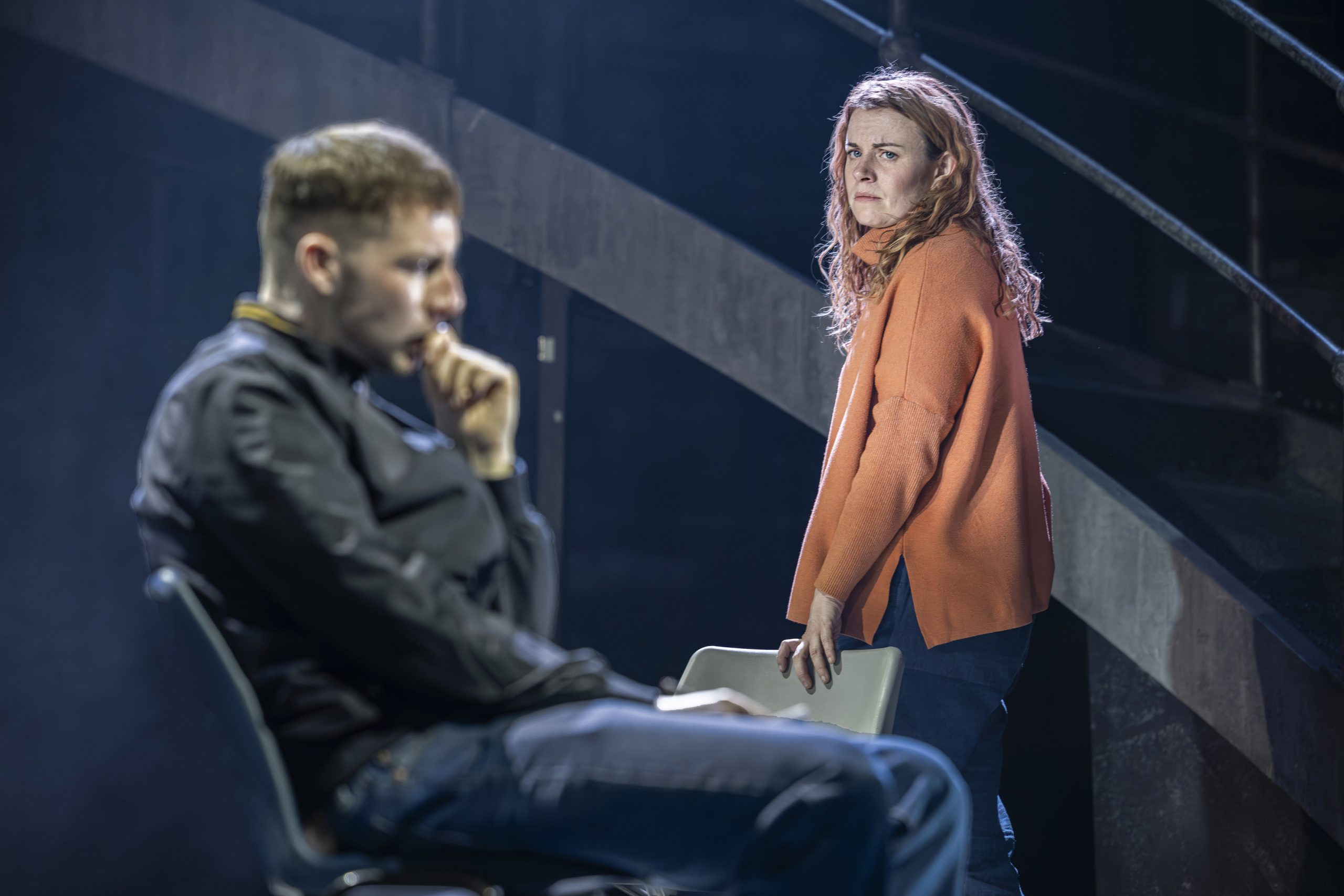 a theatre stage, one person stood in the background looking at a man sat in a chair in the front who is posing thinking