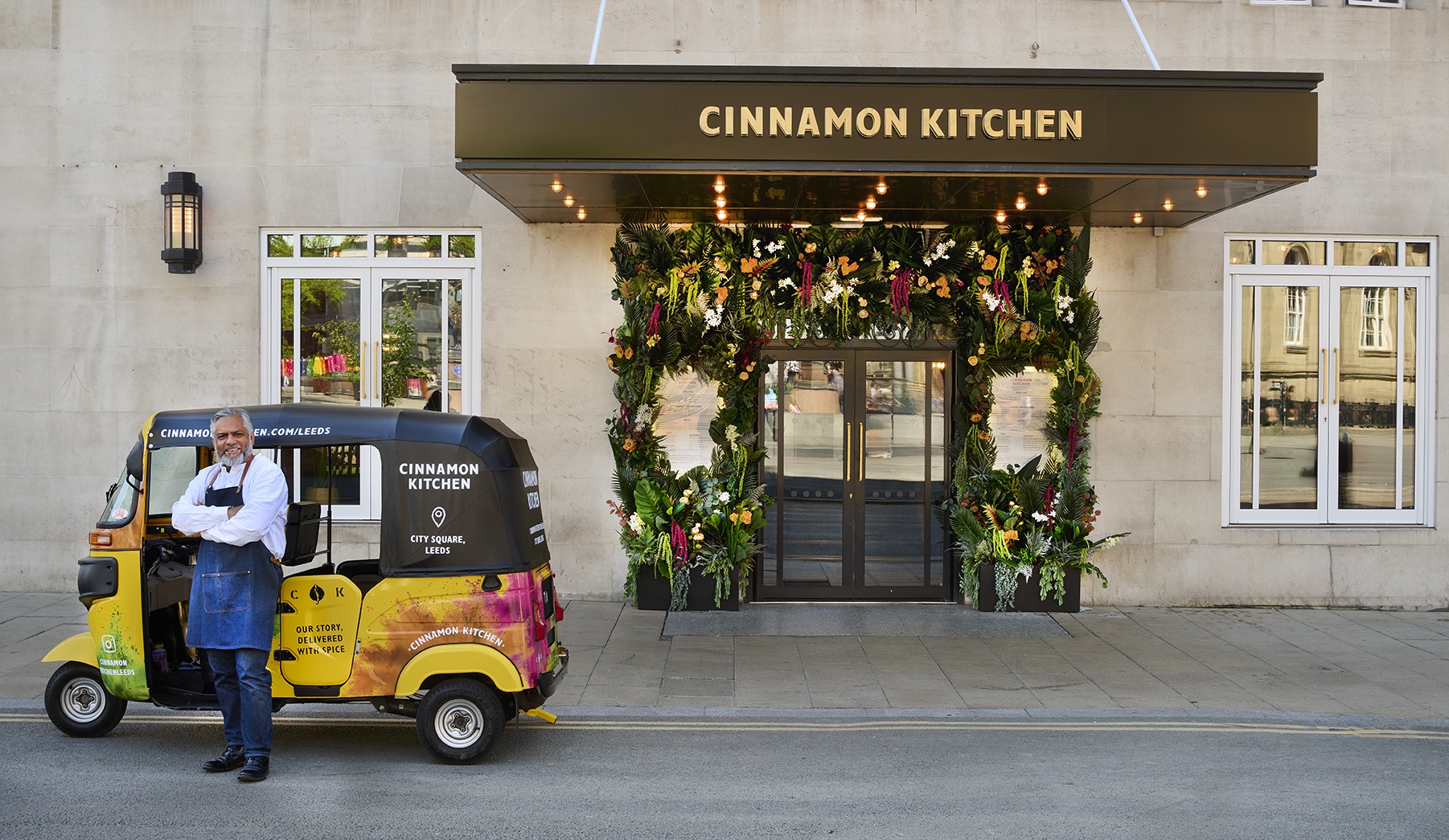 the exterior of a restaurant with a floral garland around the door, and somebody stood by a branded tuktuk vehicle outside the front door
