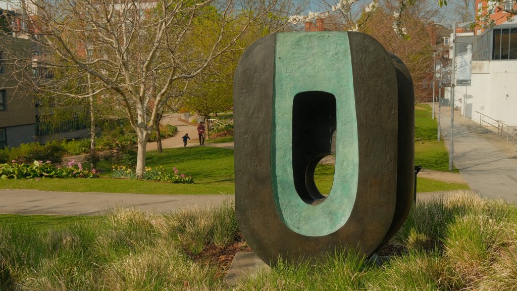an oval sculpture with a hole in the middle, sitting in a green field filled with spring flowers and blossom trees