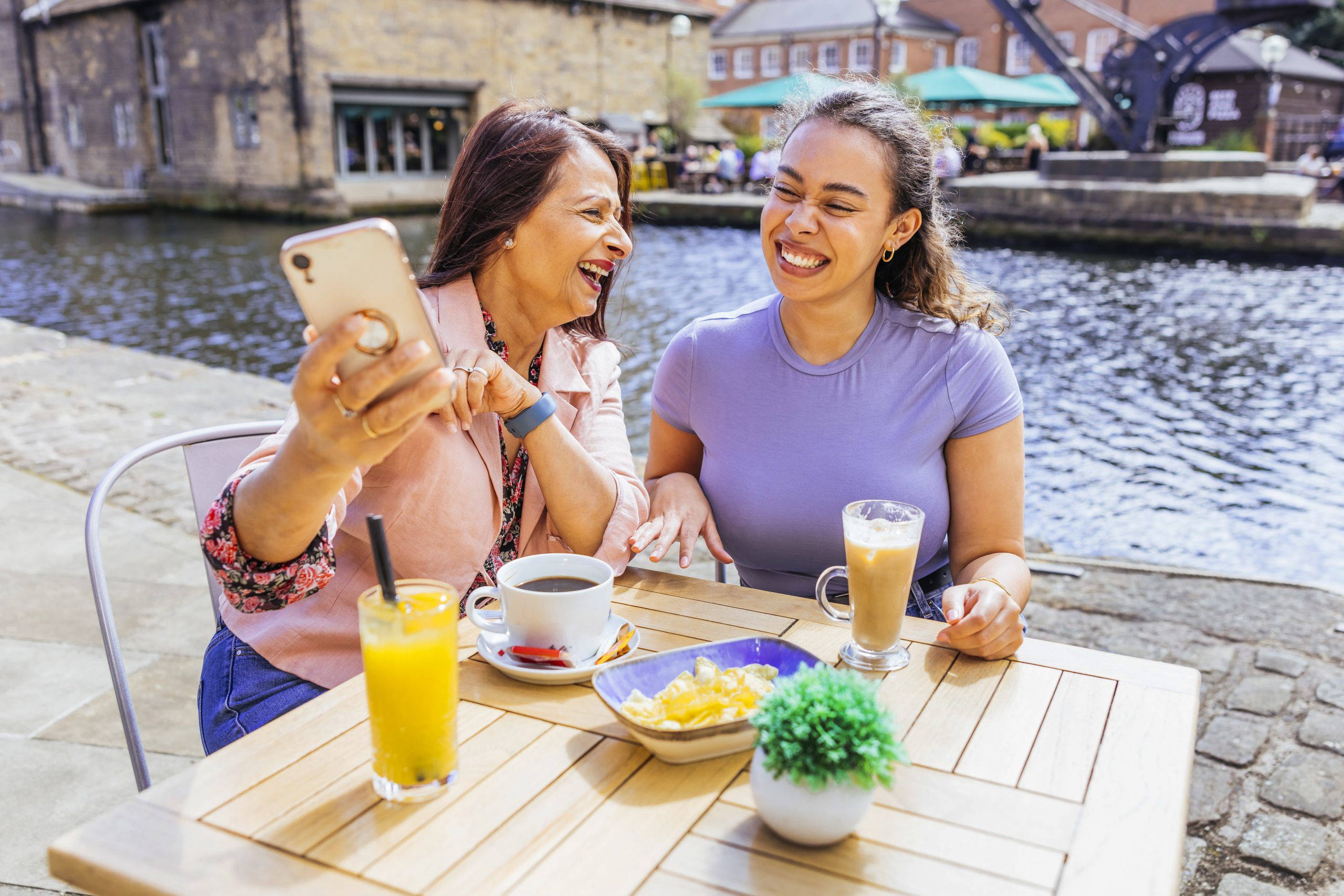 two women sat by a canalside at a table with food and drink, laughing over a phone