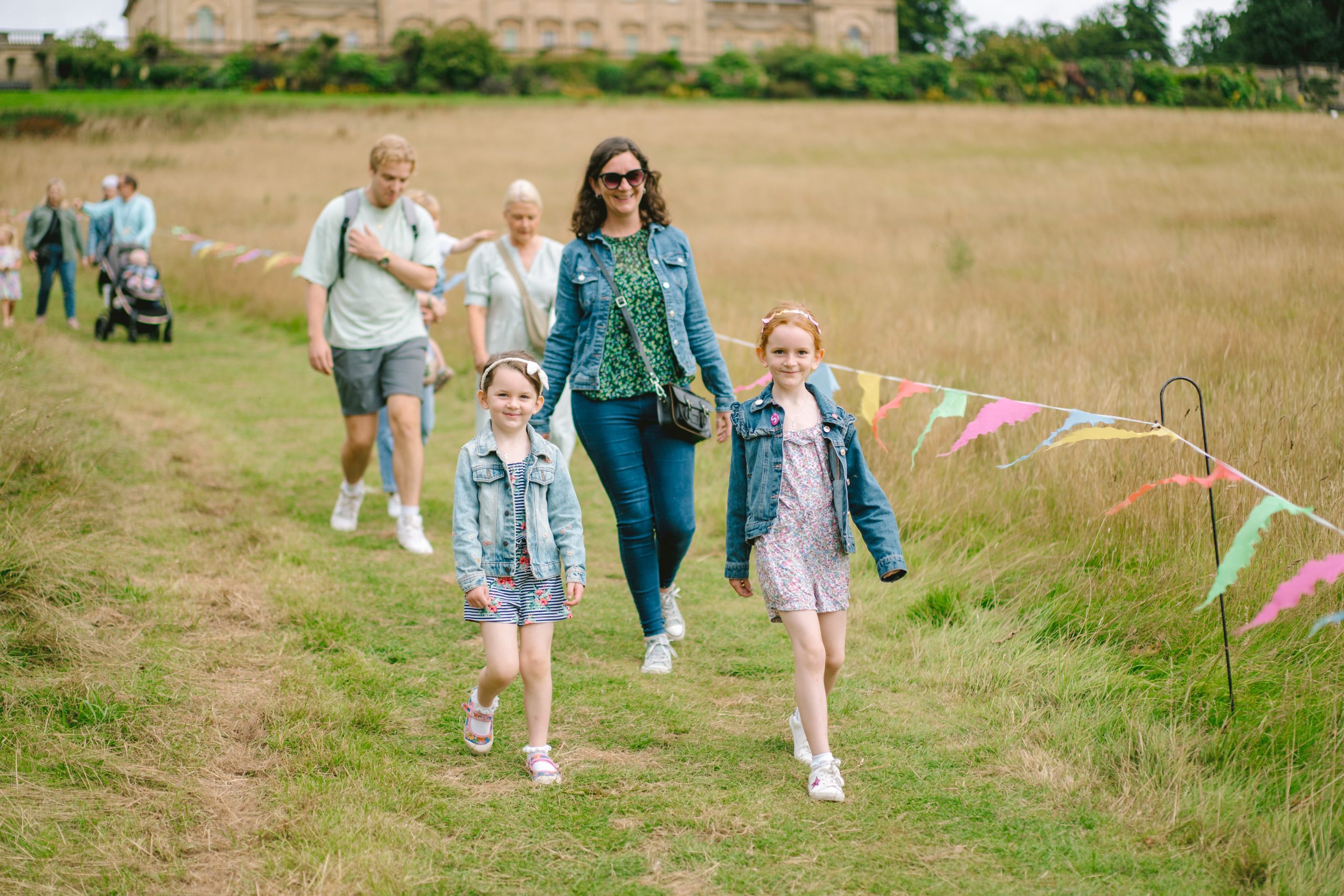 a family with children walking a field with colourful flags around them and a large historic house behind them