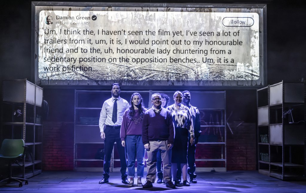 several actors on a theatre stage stood together looking at the audience whilst a tweet is projected above them saying 'from damian green - um, i think the, i haven't seen the film yet, i've seen a lot of trailers from it, um, it is, i would point out to my honourable friend and to the, uh, honourable lady chuntering from a sedentary position on the opposition benches.. um, it is a work of fiction.