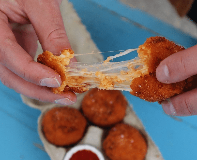 a tray of food and a dipping sauce with somebody holding a cheesy arancini bite and pulling it 