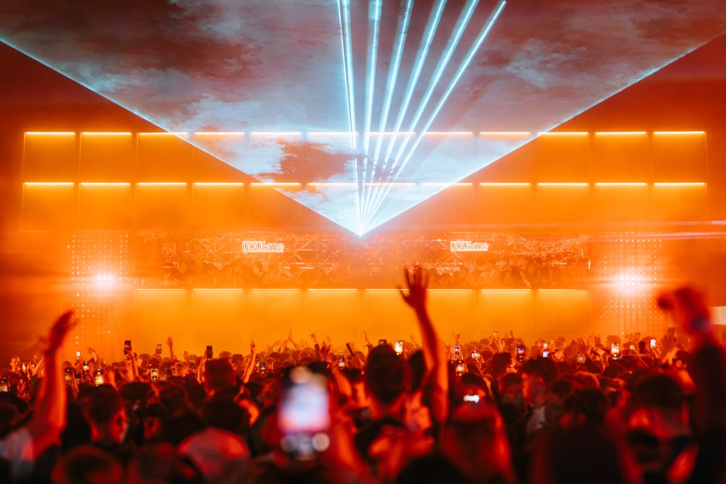 a large crowd of people a music festival with a dj in front and light beams from the stage