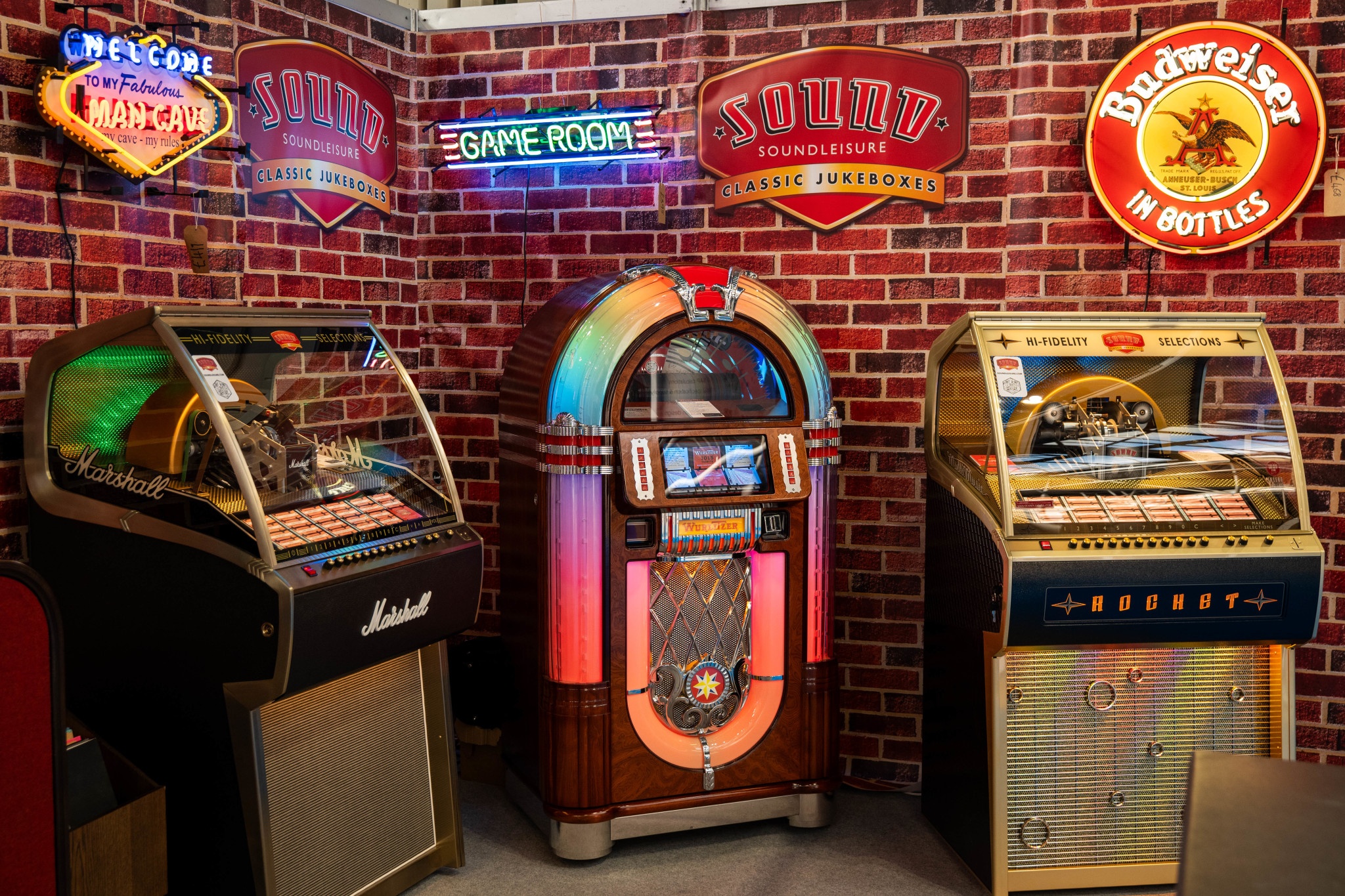several colourful jukeboxes lined up against a brick wall with neon lights across the wall