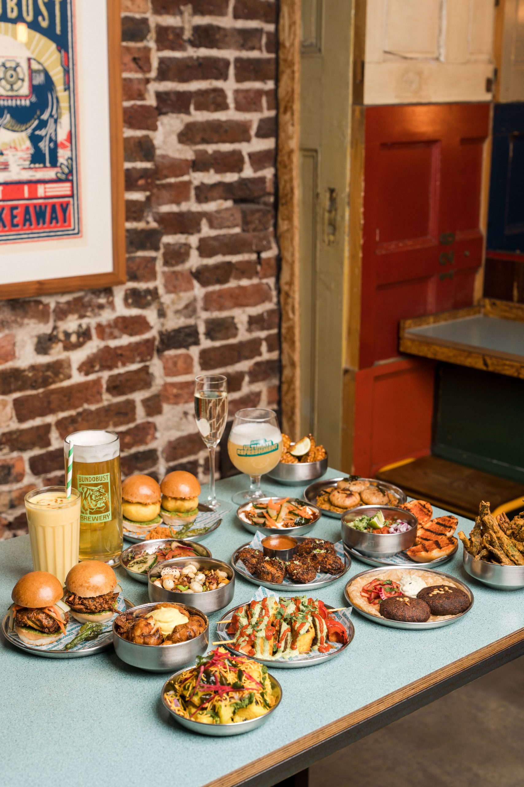 a table in front of a brick wall with several plates and drinks including burgers and beer