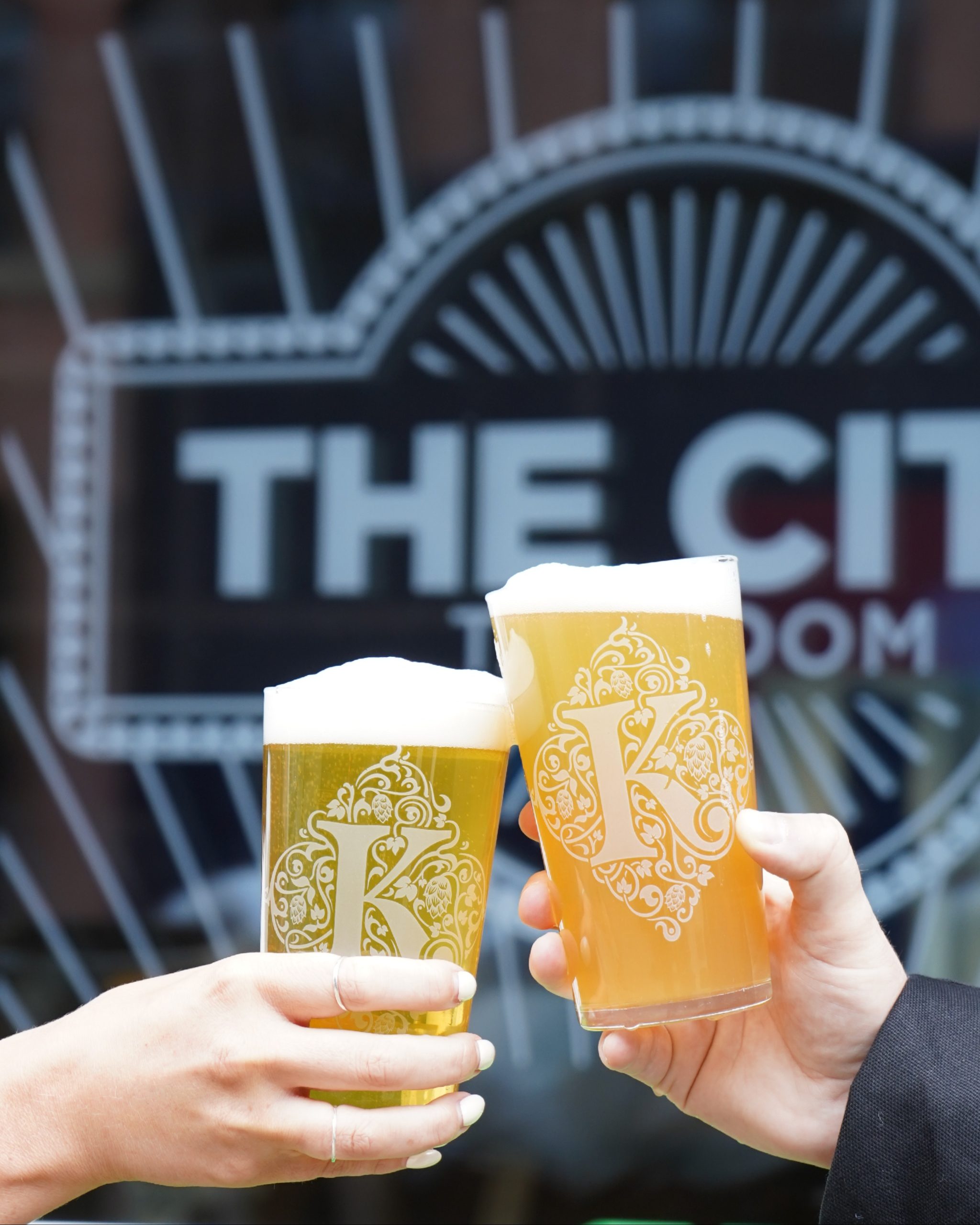 two hands clinking two pint glasses filled with beer together in front of a sign saying the city taproom