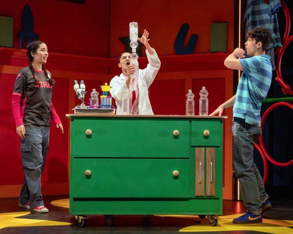 a production image of a theatre show where three actors are stood around a table with science bottles on top doing an experiment