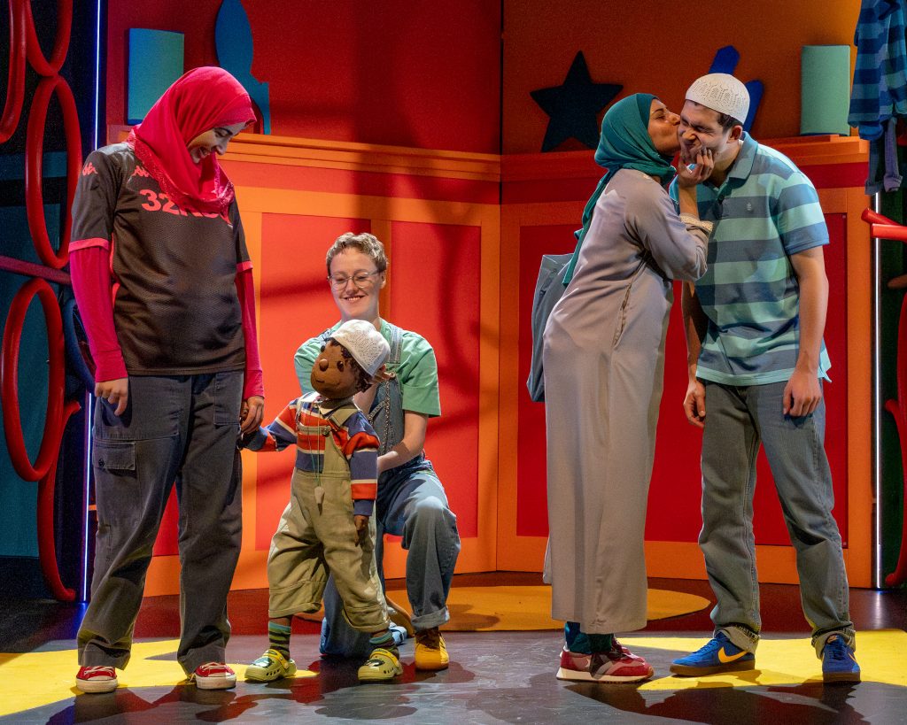 a production image of a theatre show with a handful of actors ofr varied ages with headscarves and hats on are all talking to each other