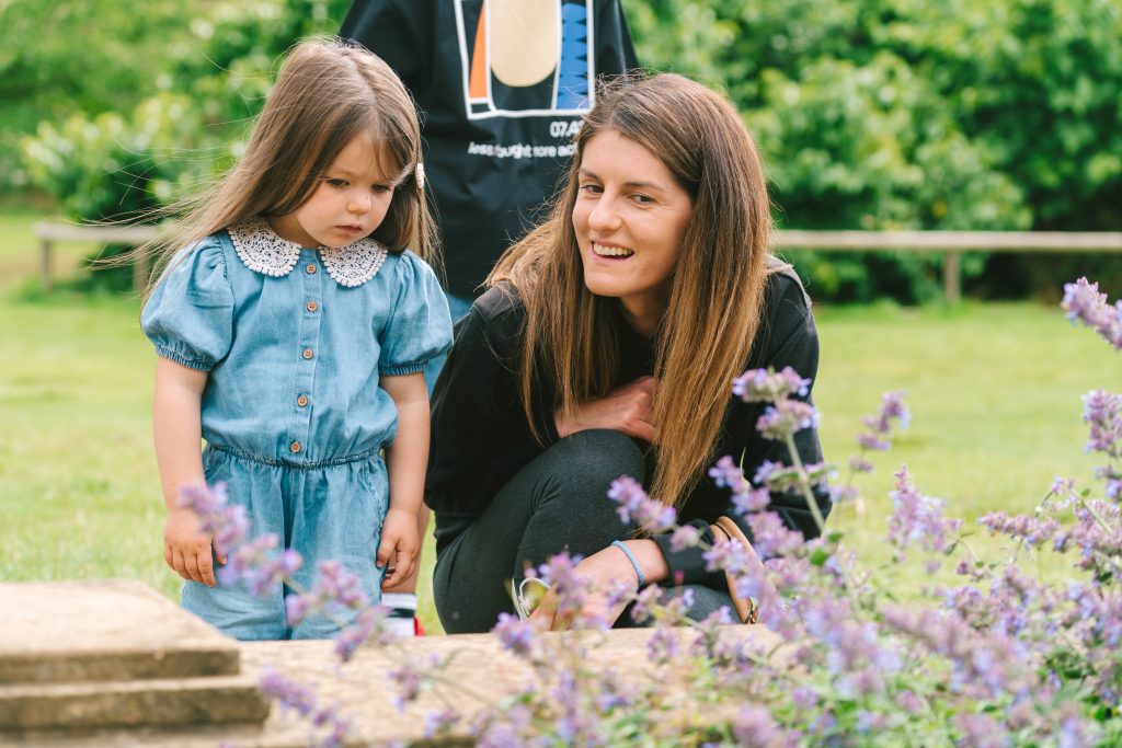 a woman kneeling down with a small child in a blue dress looking at flowers in a garden