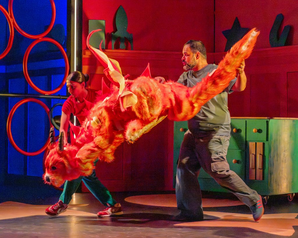 a production image of a theatre show of a colourful creature flying in between a couple of actors