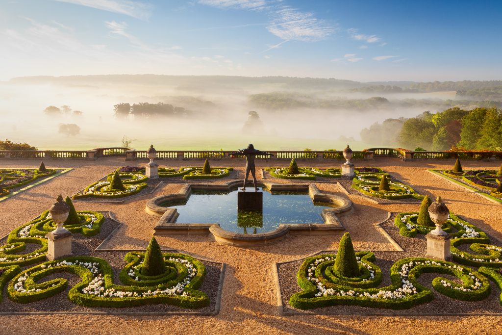 a misty morning view of a garden terrace of stately home with manicures bushes and plants, a statue in a fountain woodland in the distance
