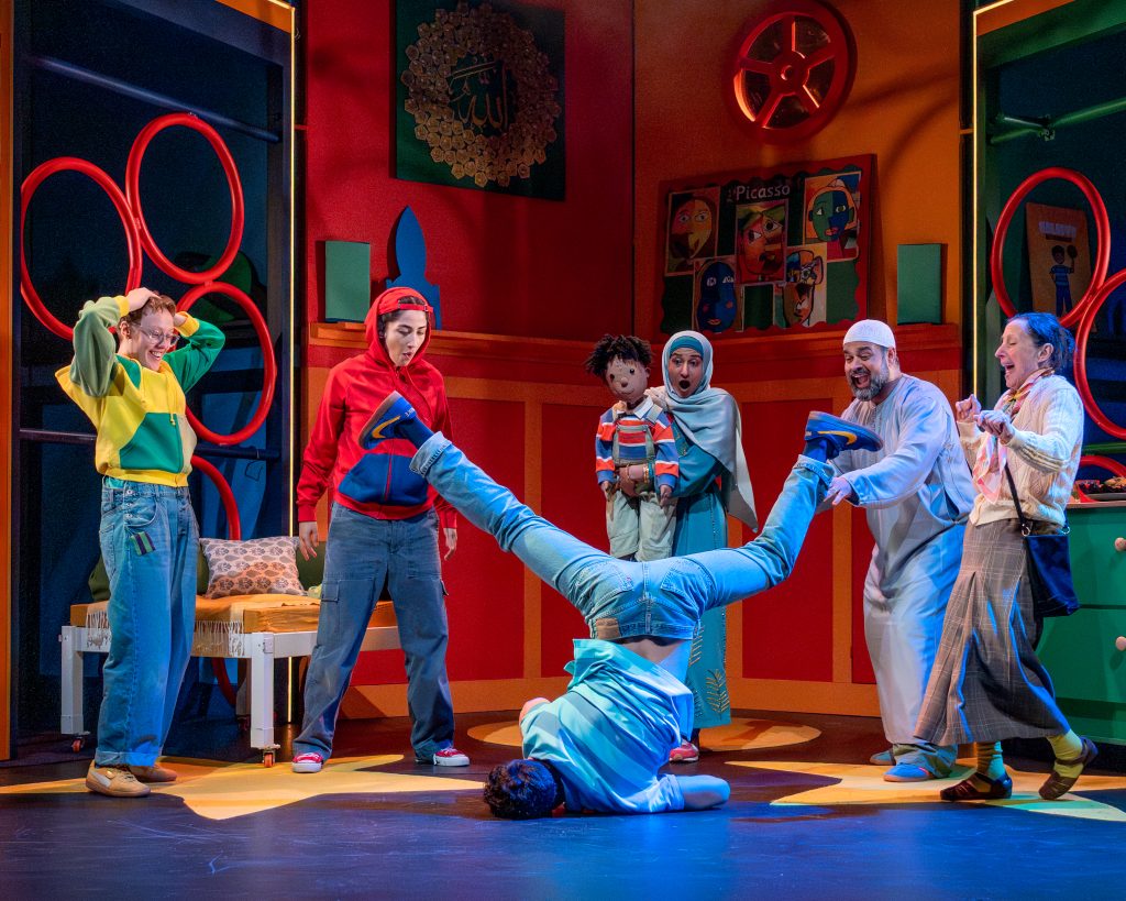 a group of actors on a colourful stage set with one breakdancing in the middle