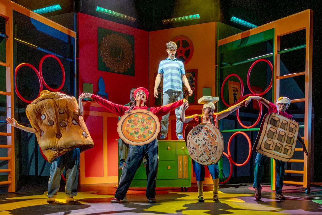 a group of actors on a colourful stage set, with one young boy stood on a table and the others with chefs hat and pizzas on them