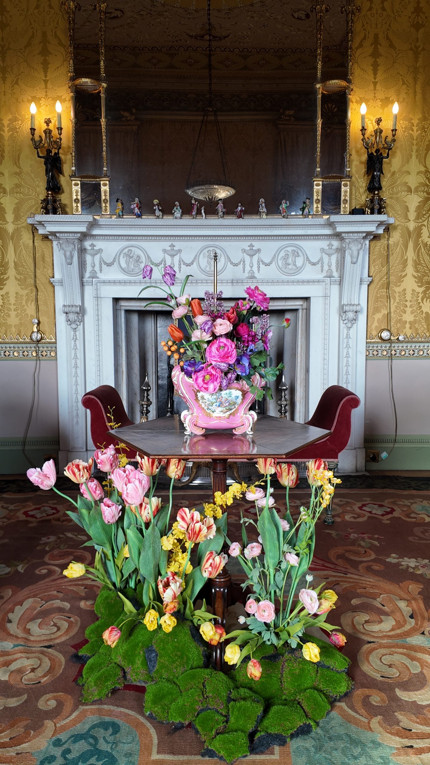 an old ornate fireplace and wooden table in front of it, surrounded by pink and yellow fresh flowers