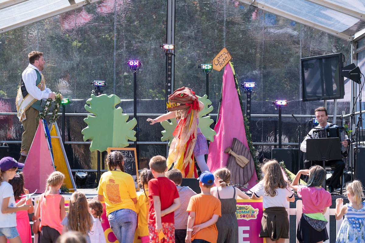 a group of children outdoors watching a performer in a colourful outfit on a stage