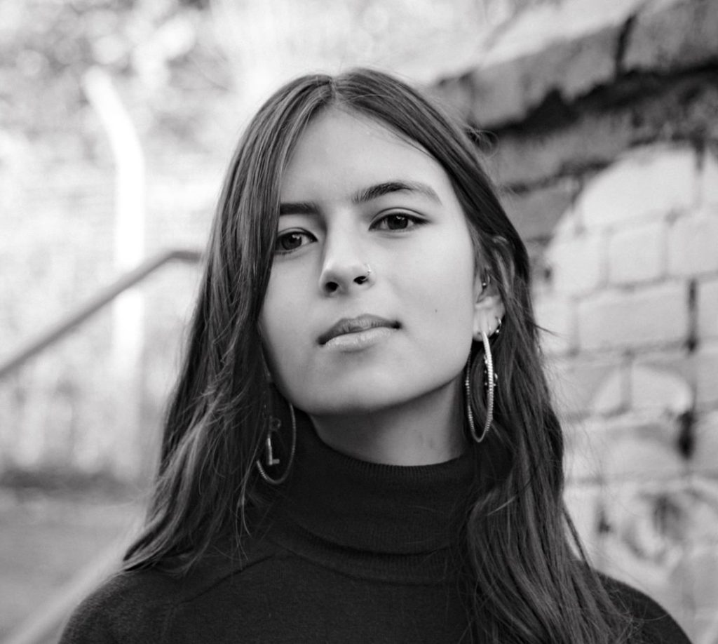 a woman with long hair, large hoop earrings and a polo neck jumper in front of a brick wall looking at the camera