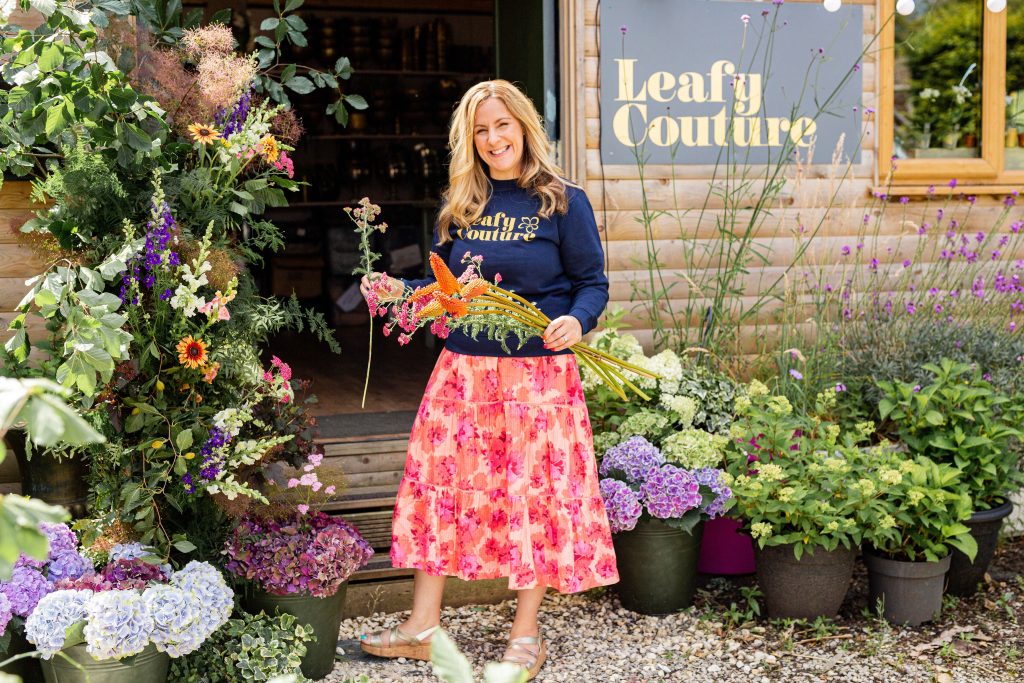 a woman standing amongst a garden filled with flowers and a sign on a brick wall behind saying leafy couture