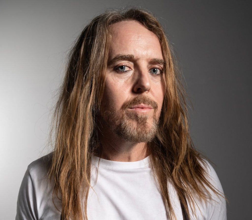 a man with long hair, a beard and a white tshirt looking at the camera
