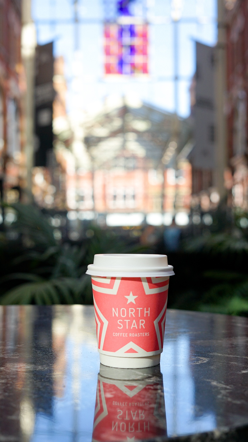 a coffee cup on a marble table, reflected in the table, inside a large shopping mall with a stained glass roof and greenery through the bottom
