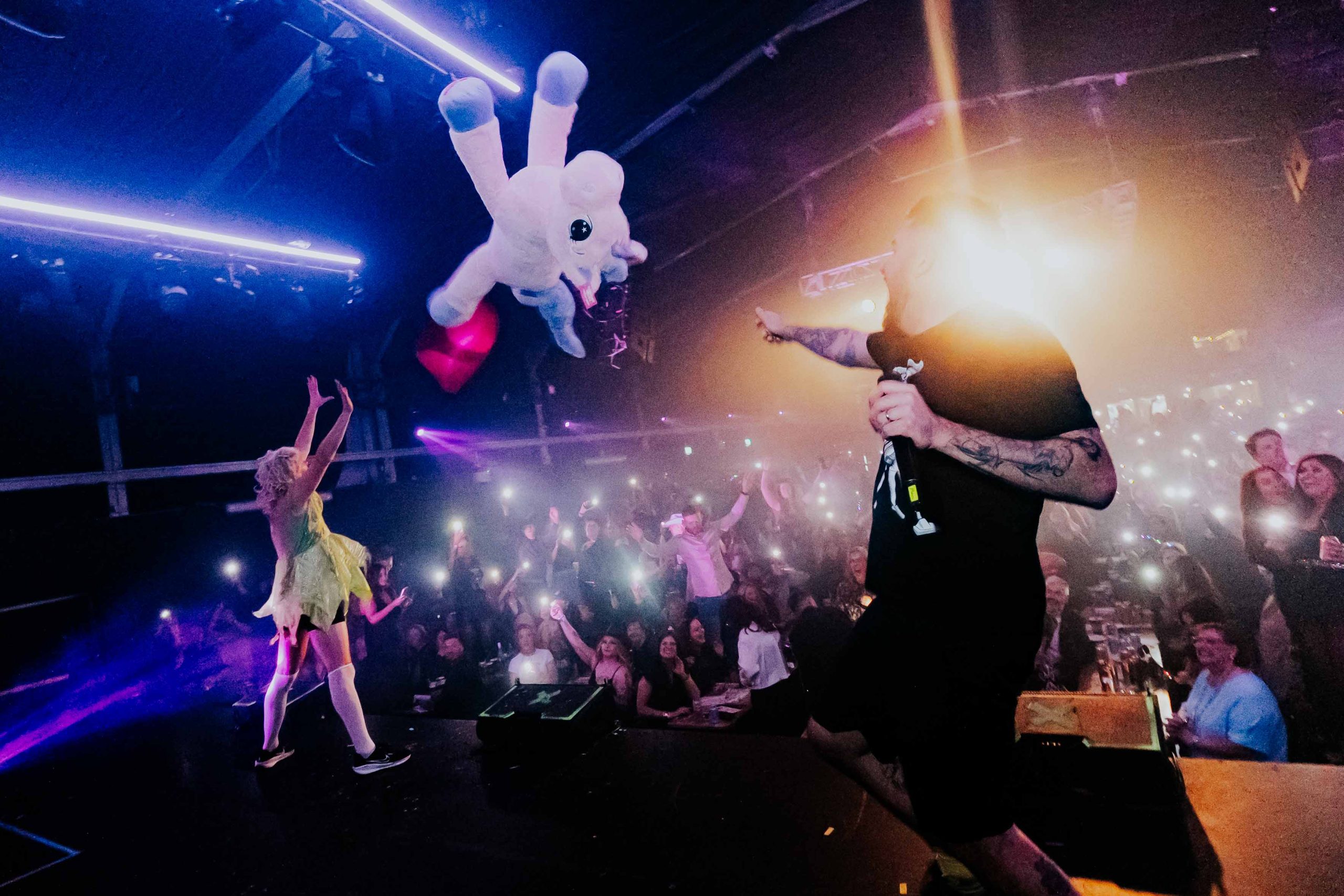a large venue with a large crowd of people whilst people perform on a stage throwing a large unicorn plush into the air