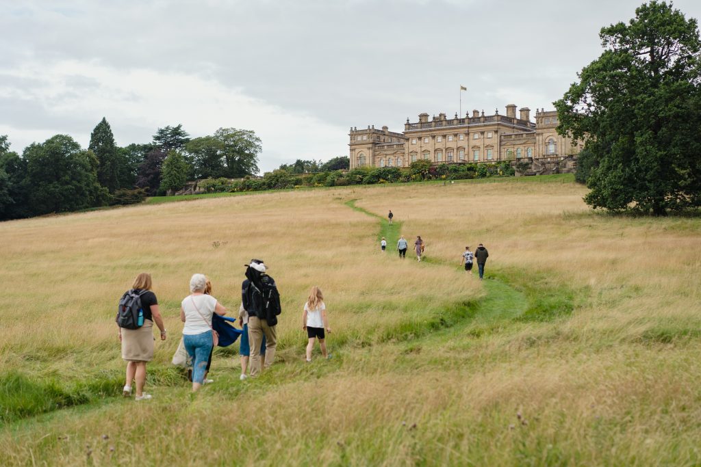 a large field with a historic stately home at the top of a hill with people walking through a trail made in the grass