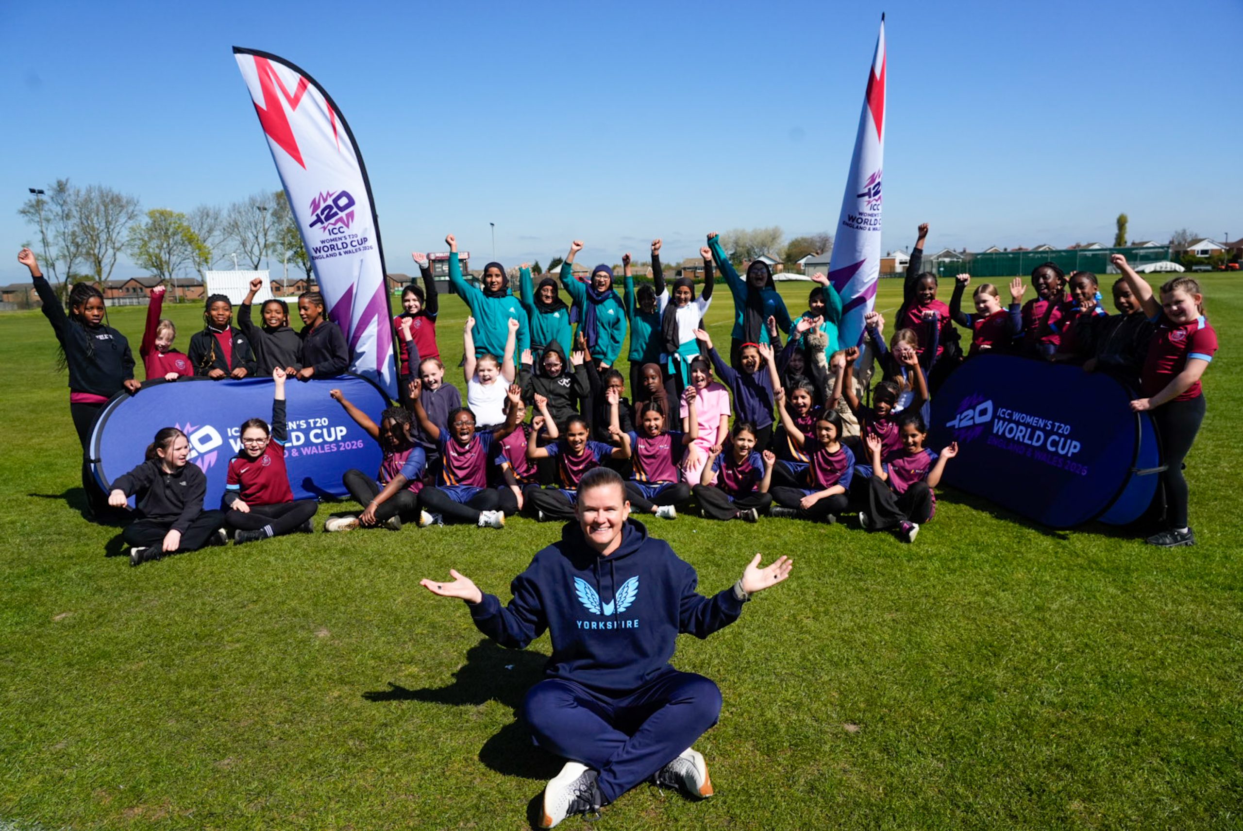 a field with a woman sat cross legged in front of a crowd of young girls all cheering