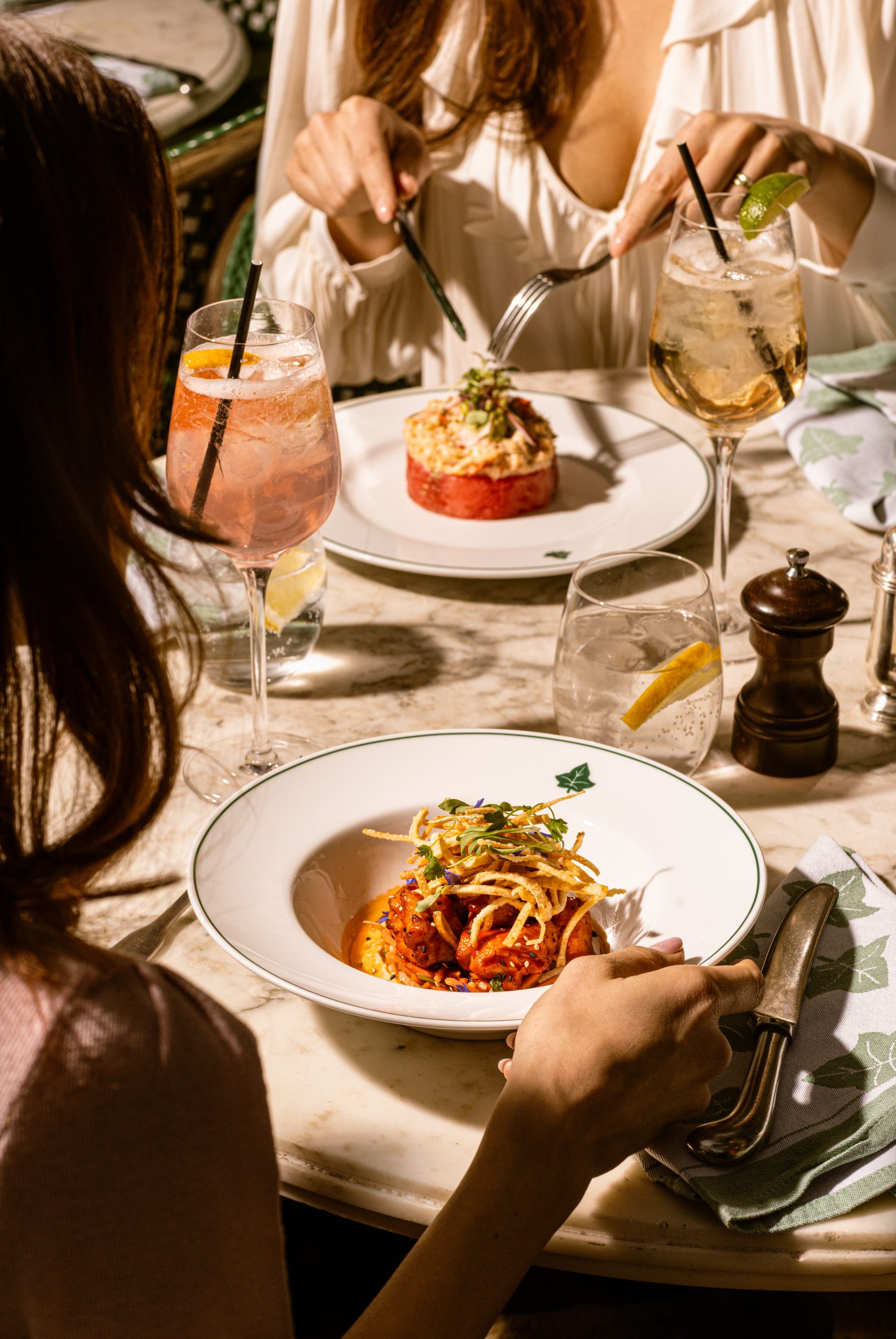 a couple of people sat a table with plates of food in front of the, salt and pepper shakers and some cocktail glasses