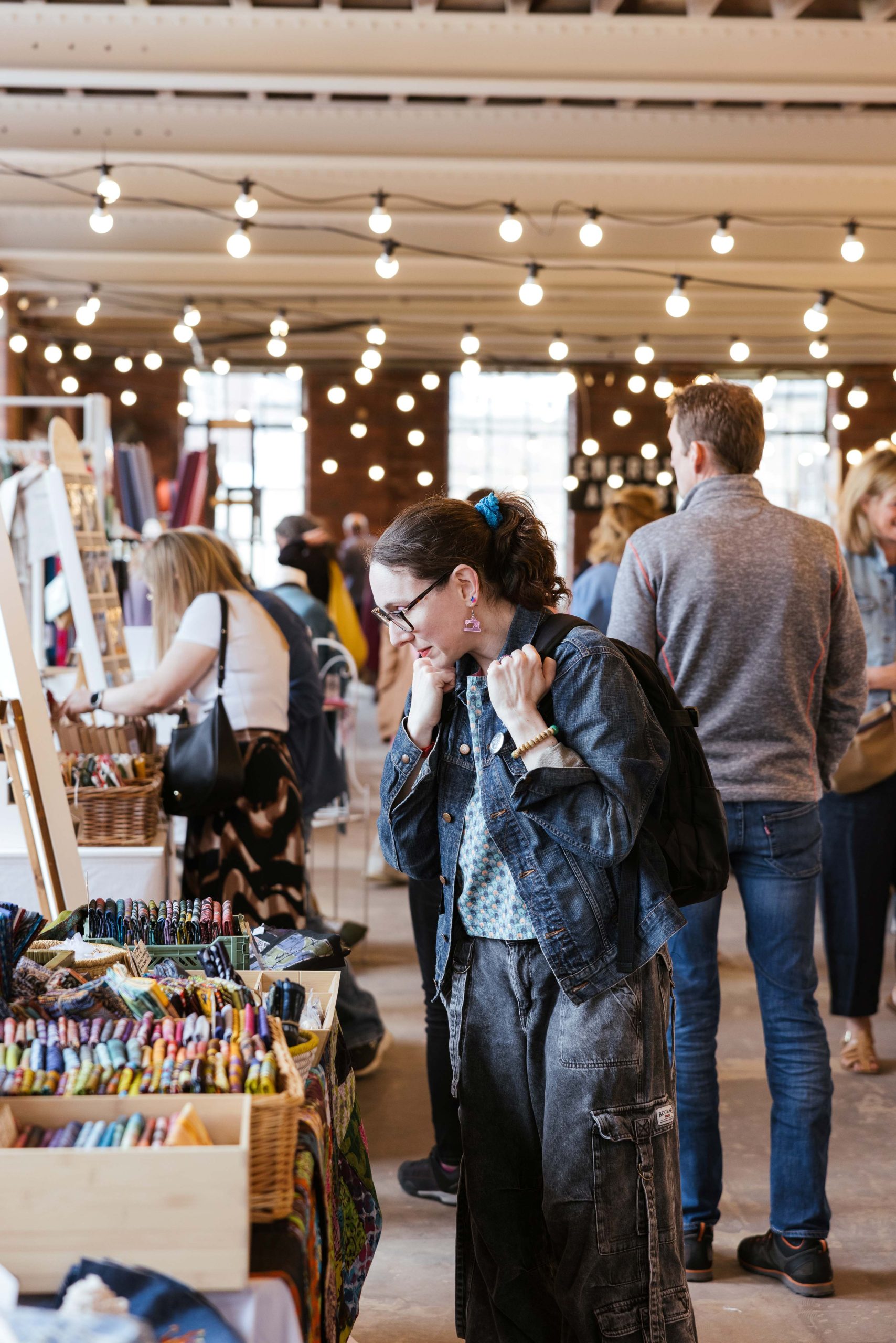 a brick room with fairy lights strung up on the ceiling and people walking around looking at tables filled with market stall crafts