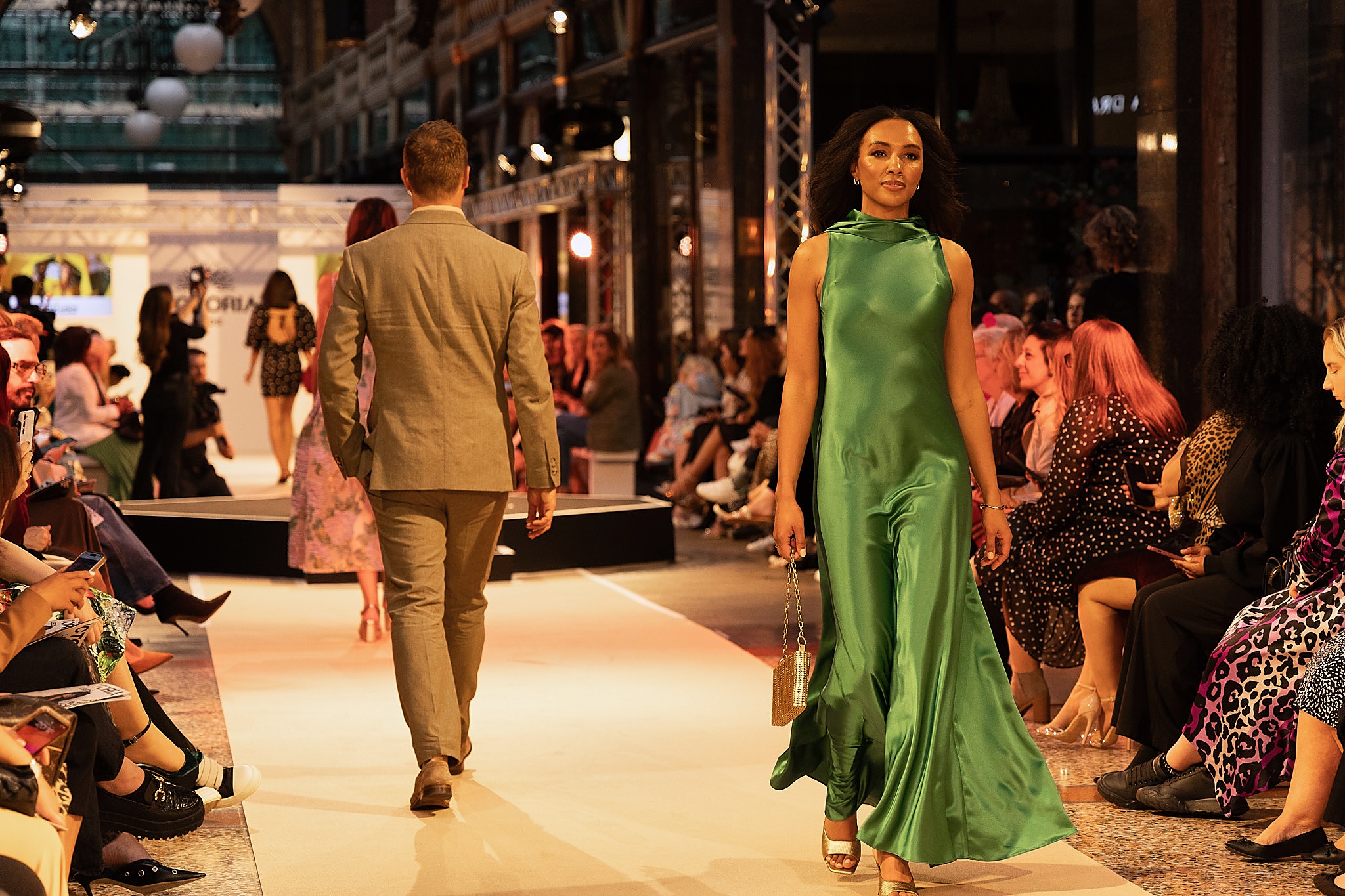 a woman in a long green gown walking on a runway with people watching and a man in a suit walking down the opposite side of the runway