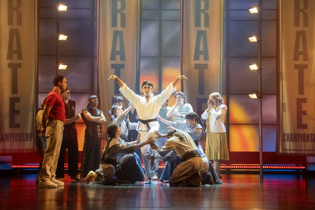 a theatre stage with a troupe of actors in the middle with one in the spotlight wearing karate white outfit