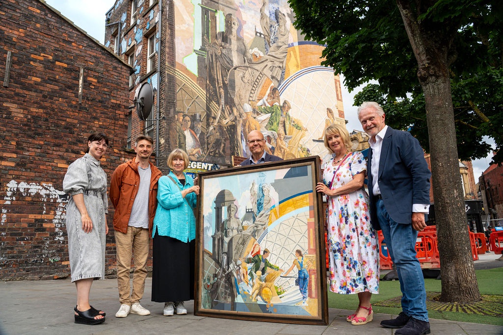 six people stood together with a painting in a frame in front of them of the mural behind them on the side of a building
